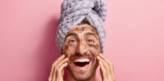 Male beauty concept. Happy joyous man applies coffee scrub on face, removes dark dotes, wants to look refreshed, has wrapped towel on head, poses against pink background topless, shows white teeth