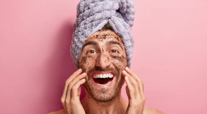 Male beauty concept. Happy joyous man applies coffee scrub on face, removes dark dotes, wants to look refreshed, has wrapped towel on head, poses against pink background topless, shows white teeth